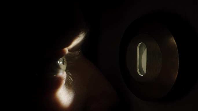 一个男人往门锁里偷看。锁眼里的光线照在他的眼睛里，近景