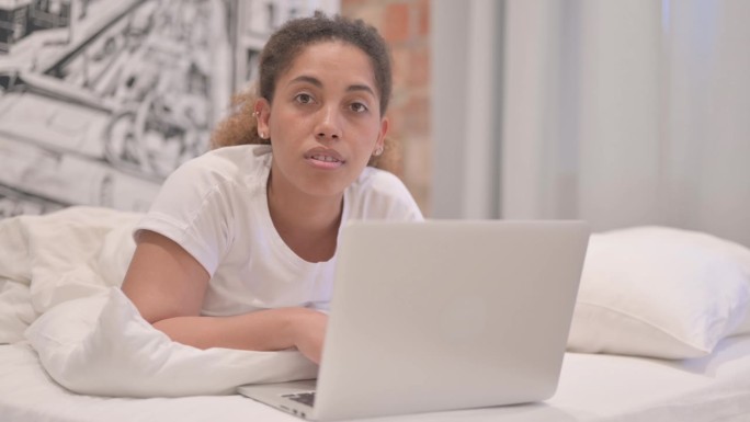 一名非洲裔美国女性趴在床上，一边用笔记本电脑工作，一边对着镜头微笑