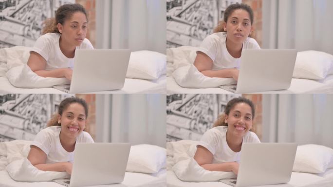 一名非洲裔美国女性趴在床上，一边用笔记本电脑工作，一边对着镜头微笑