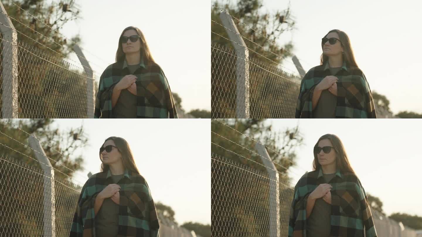 一名身穿格子衬衫的年轻女子沿着装有倒刺铁丝的铁丝网走着。日落时分，在森林附近。