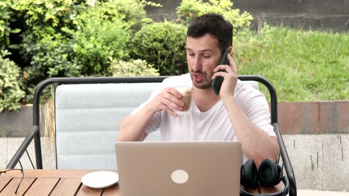 在户外咖啡馆，拿着一杯咖啡在笔记本电脑上工作的男人。坐在街边咖啡馆的桌边使用电脑的商人的特写肖像。