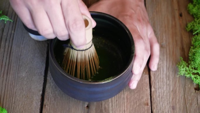 咖啡师用木勺将有机抹茶绿茶粉倒入碗中。抹茶绿茶的制备过程在一个黑色的碗在木桌背景。一杯抹茶绿茶放在棕