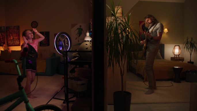 两个房间被墙隔开。男人弹吉他，小女孩录舞蹈录影带