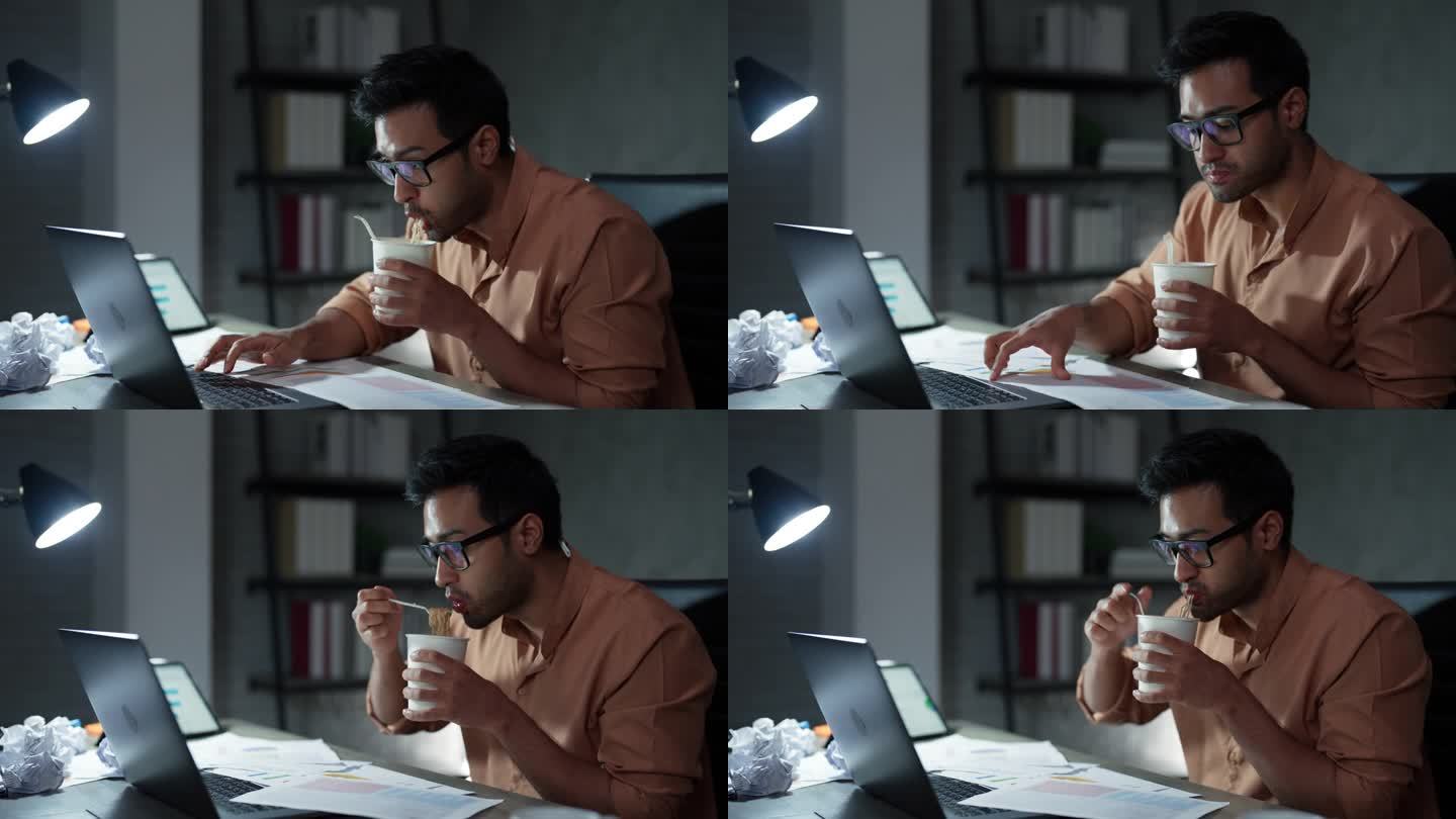 一名亚洲年轻男子在加班期间一边做项目计划，一边坐在办公桌前吃泡面。