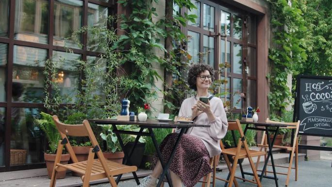 快乐的女人坐在咖啡馆露台上，手里拿着智能手机