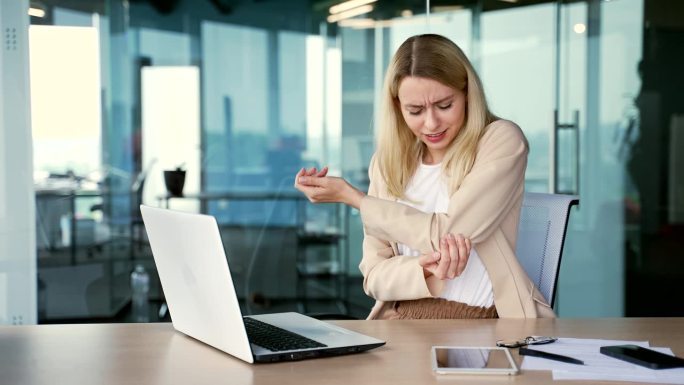疲惫的年轻女商人在现代办公室坐在办公桌前用笔记本电脑工作时肘部疼痛。