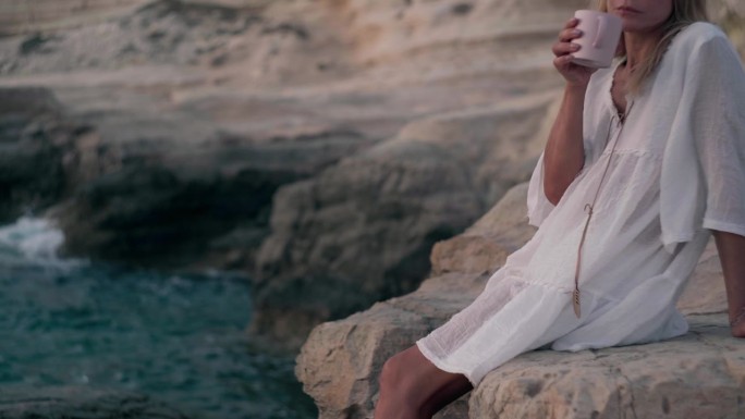 右边是一个无法辨认的女人，她拿着茶杯坐在地中海的岩石上。高加索女游客端着咖啡在塞浦路斯欣赏日落。