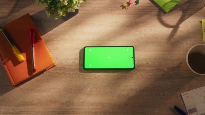 一个智能手机与模拟绿色屏幕显示的自顶向下视图。一个手机水平躺在一个创意办公室的木桌上。数字营销和屏幕