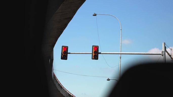 从汽车上看十字路口桥下的柱子上挂着两个交通灯。红色的光和蓝色的天空为背景