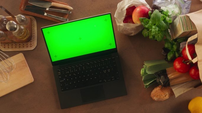 一台笔记本电脑的俯视图，配有模拟绿屏Chromakey显示屏和运动追踪器，它站在厨房的木桌上，旁边是