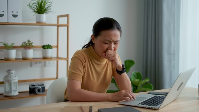 一名在手提电脑前工作的亚洲妇女发烧、流鼻涕、鼻塞和喉咙痛。