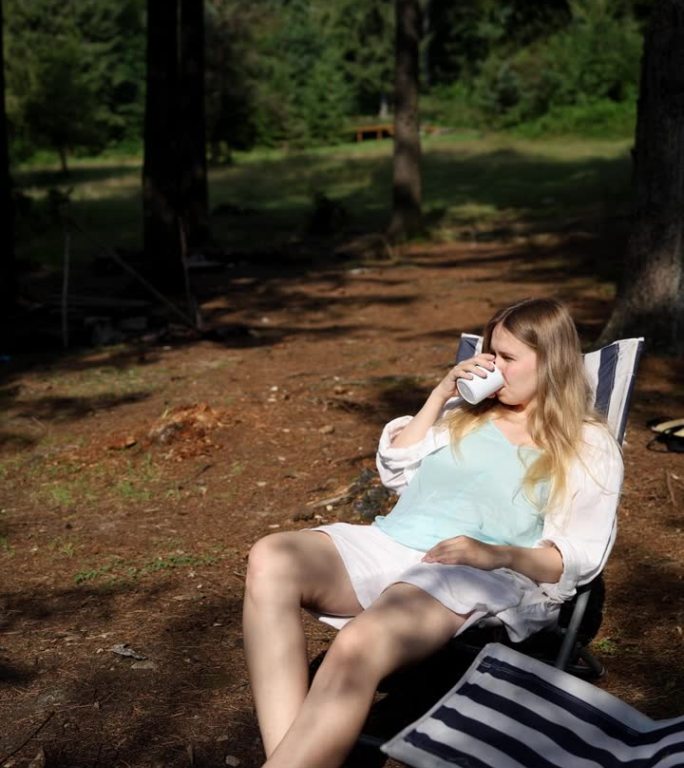 美丽的女孩穿着夏天的衣服，躺在自然的躺椅上，在高山中喝着白色的杯子里的咖啡，背景是一片高大的森林。垂