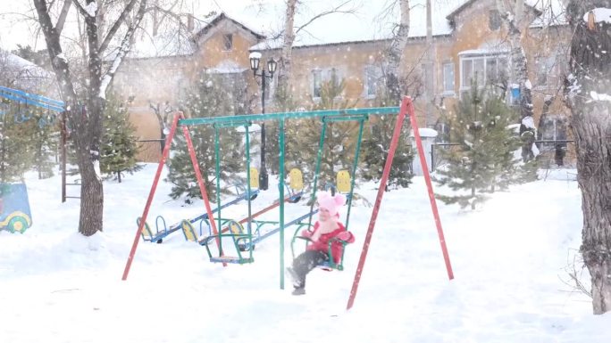 冬天，蹒跚学步的小女孩在操场上玩耍。下雪的一天。下雪后，孩子在操场上独自荡秋千。