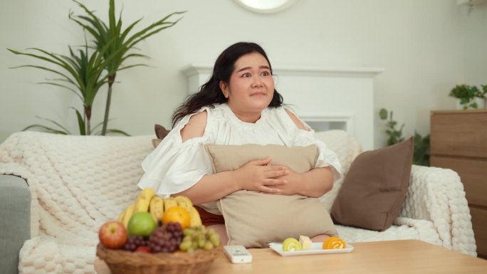 一个肥胖的亚洲年轻女子坐在沙发上，拿着遥控器换频道，看一部爱情剧。