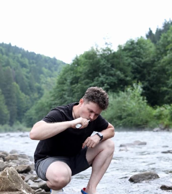 一个男孩游客坐在山上河边的石头上，用电子牙刷刷牙。在山上洗澡。垂直视频