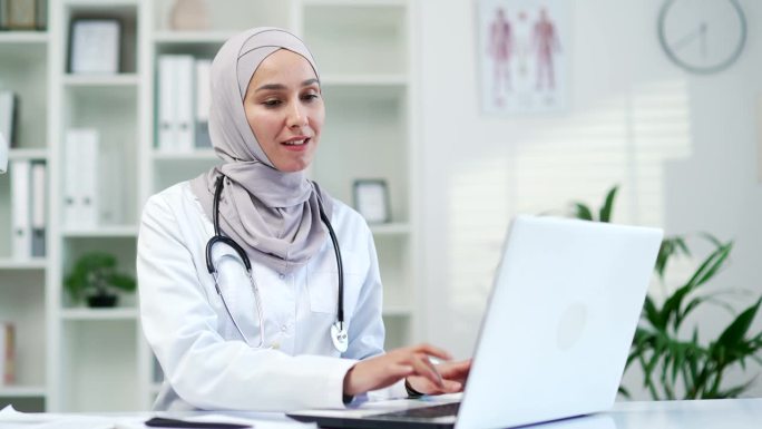 穆斯林女医生在诊所用笔记本电脑打字，计划研究，阅读在线报告。