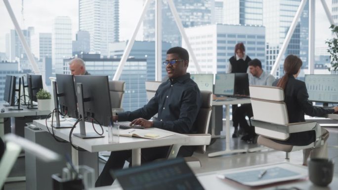 非洲裔美国男性数据分析师在不同公司办公室的台式电脑上打字。与同事一起在咨询公司工作的专业黑人。