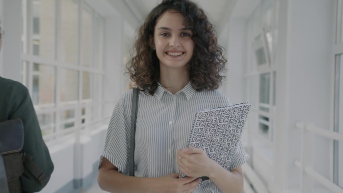 一个快乐的女孩站在现代大学的大厅里，拿着教科书，微笑着看着学生们走过