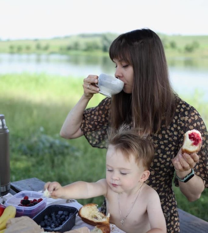 妈妈和小儿子在大自然中喝茶吃糖。女人和孩子拿着牛角面包，以湖为背景。垂直视频