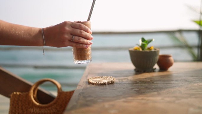 户外热带海景咖啡厅，女服务员将装有鸡尾酒和金属吸管的高脚杯放在木桌上。将冰咖啡或冰沙放在观景屋顶餐厅