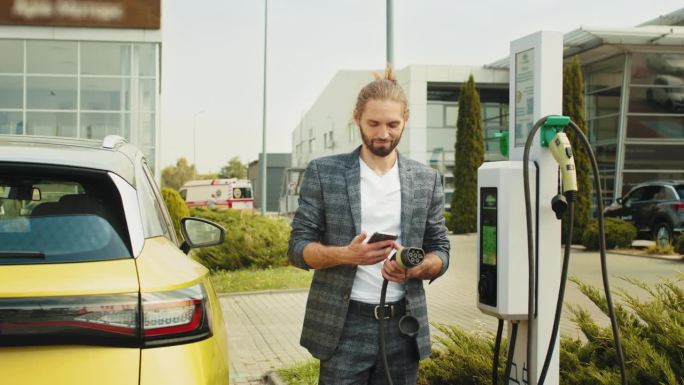 商人拿着智能手机在电动汽车充电站给汽车充电，特写。男子手持智能手机在电动汽车充电站为汽车充电，特写