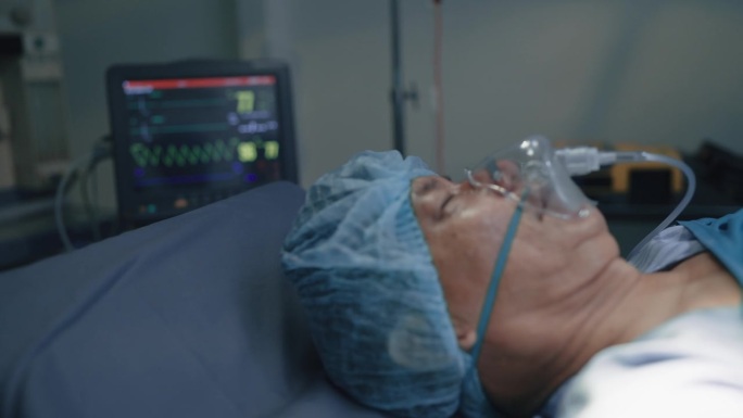 佩戴医用呼吸机躺在手术室的手术床上，近距离观察患者面部。模糊的背景显示病人的心率