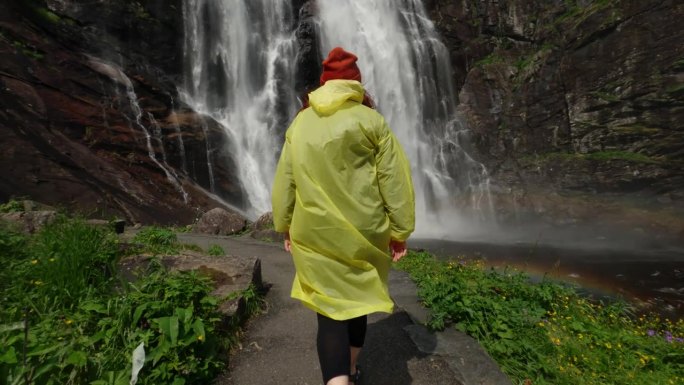 穿着黄色雨衣的年轻女孩徒步旅行者走到高地的瀑布前，举起双手，享受自然和生活。在山中旅行，在旅途中冒险