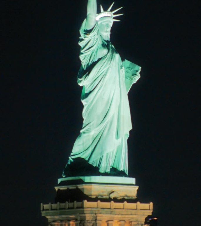 垂直屏幕:环绕自由女神像的空中飞行。直升机拍摄的美丽夜晚的爱国纪念碑。带着灯光的渡船载着游客往返于曼