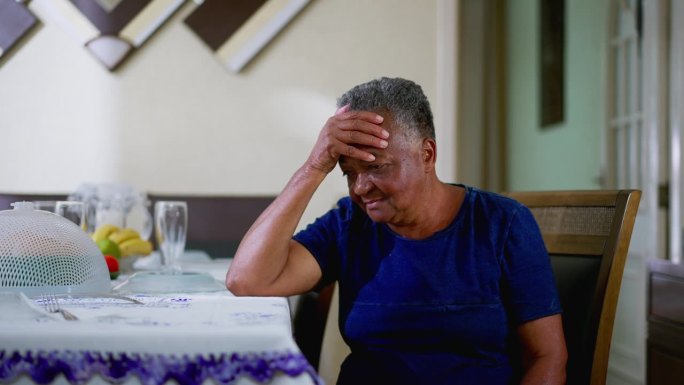 一位心事重重的黑人老年妇女独自坐在家里，手捂着额头，患有精神焦虑症。一位忧心忡忡的非裔美国老妇人