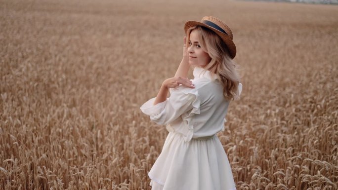 一个年轻的金发女孩戴着草帽，在阳光明媚的日子里走在金色的麦田里，带着迷人的微笑转向镜头