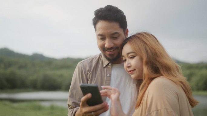 一对亚洲夫妇在大自然中散步，站在草地上，用智能手机自拍，并在社交媒体上重新查看照片。