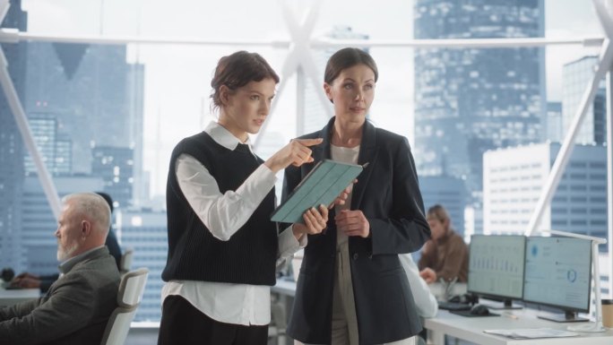 职业女性首席执行官和她的助理站在市中心现代多样的摩天大楼办公室里。两名白人女性在平板电脑上完成金融科