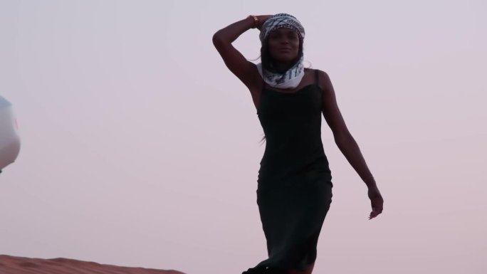 一位身穿黑色长裙、围着阿拉伯头巾、皮肤黝黑的年轻女子赤脚走在沙漠的沙地上，向一辆停在沙丘上的白色SU