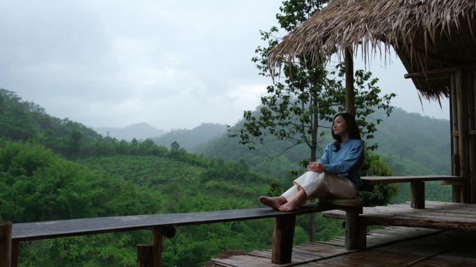 一位年轻女子坐在木阳台上，在雨中看着美丽的山景