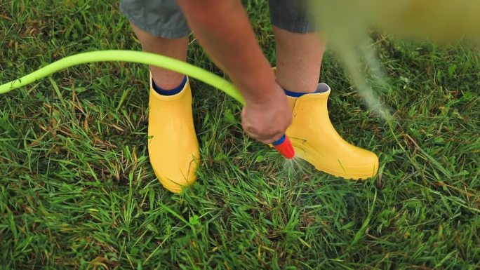 一名妇女在地里干活后，用花园水管在花园里洗黄色胶靴上的泥