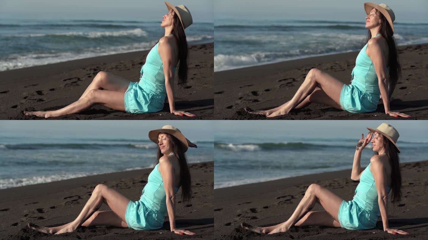 50岁的赤脚妇女坐在沙滩上，穿着短夏装，戴着草帽，放松地晒着日光浴