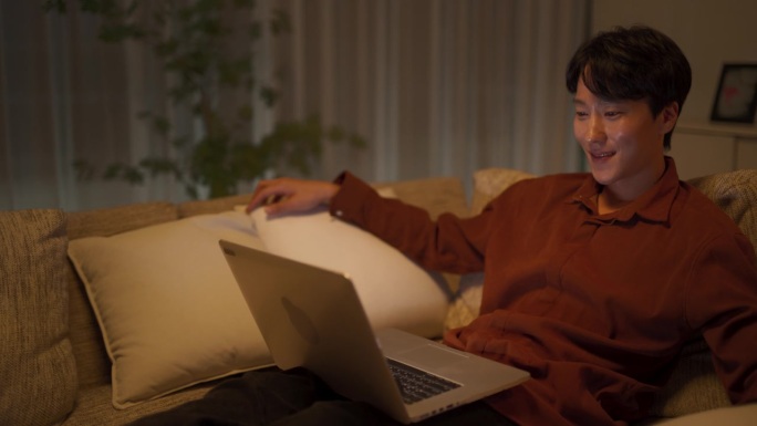 韩国帅哥躺在家里的沙发上，用笔记本电脑在网上看放松的视频。年轻人与创作者联系，留下令人鼓舞的反馈意见