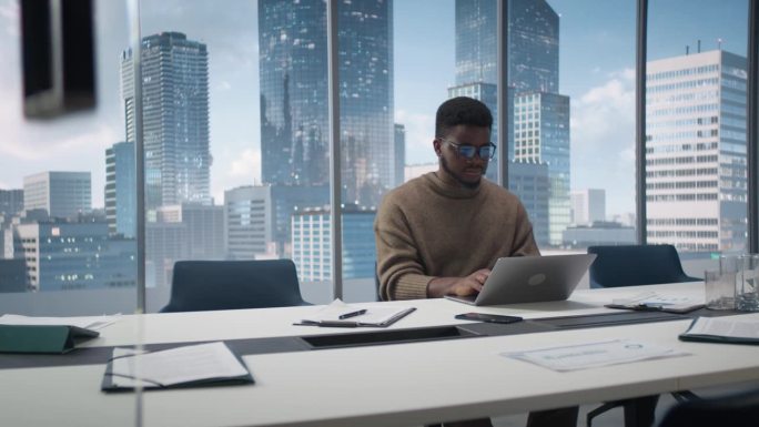 自信的黑人商人坐在现代办公室的办公桌前，使用笔记本电脑，旁边的窗户可以看到大城市的摩天大楼。专注销售