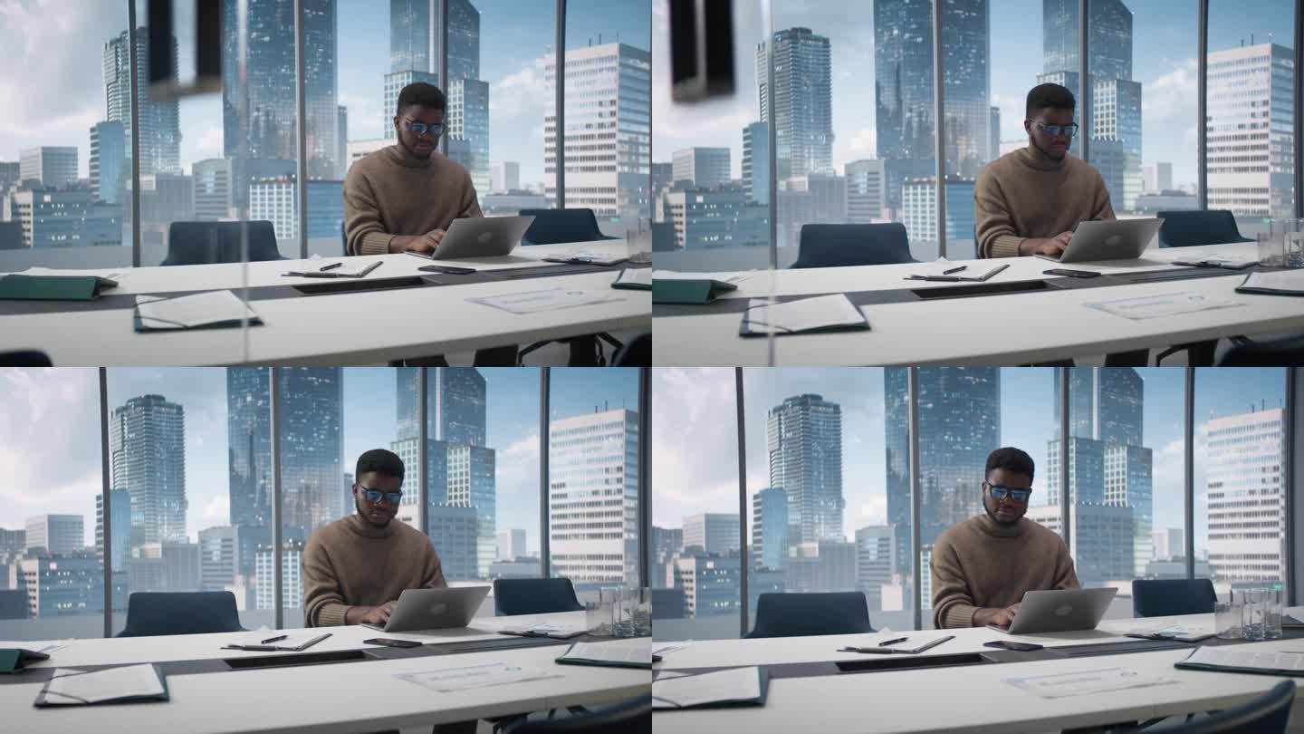 自信的黑人商人坐在现代办公室的办公桌前，使用笔记本电脑，旁边的窗户可以看到大城市的摩天大楼。专注销售