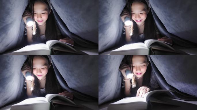 可爱的亚洲女孩晚上在卧室里拿着手电筒盖着毯子看书