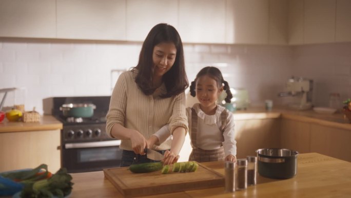 韩国妇女和女儿在厨房一起做饭的肖像。妈妈教她的小女孩如何切蔬菜。小女孩帮妈妈准备午餐。快乐的童年