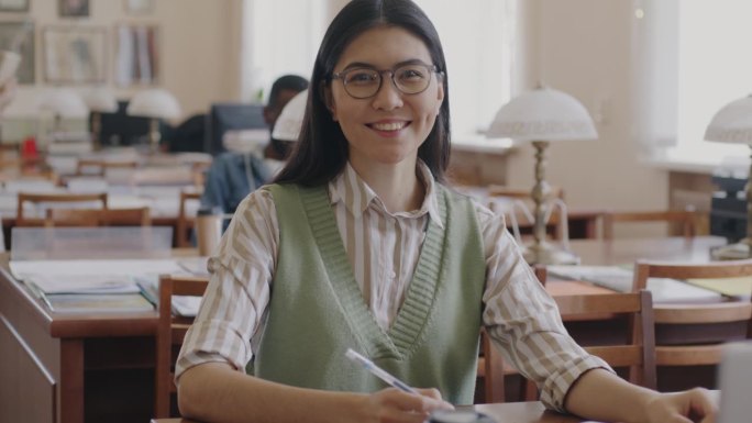 快乐的亚洲女学生的肖像，戴着眼镜，穿着休闲服，坐在大学图书馆里，拿着书，微笑着看着镜头