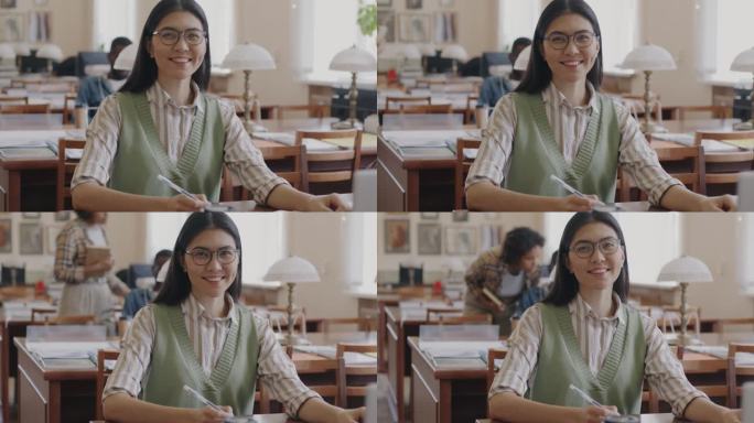 快乐的亚洲女学生的肖像，戴着眼镜，穿着休闲服，坐在大学图书馆里，拿着书，微笑着看着镜头