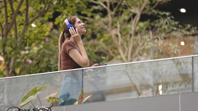 美女站在那里，戴着蓝牙耳机，愉快地听着音乐，从智能手机通过高速无线网络收听。在城市花园
