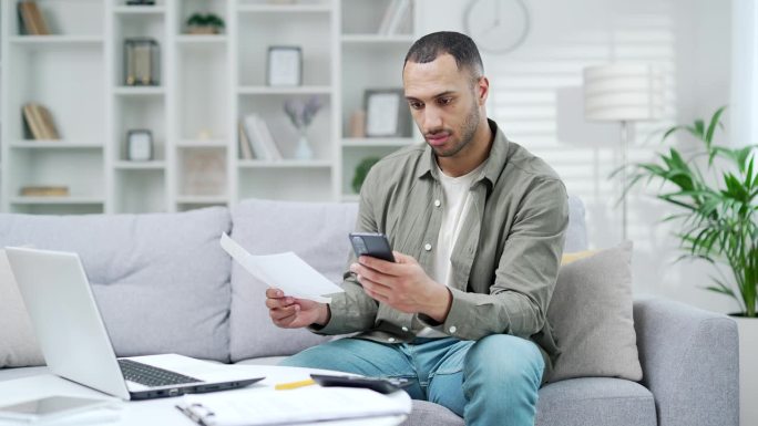 快乐的男人坐在家里的房间里，用手机上的智能手机使用手机应用程序支付账单