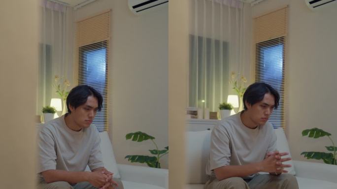 悲伤沮丧的年轻亚洲男子坐在家里客厅的沙发上。生活方式花时间的房子。垂直的屏幕。
