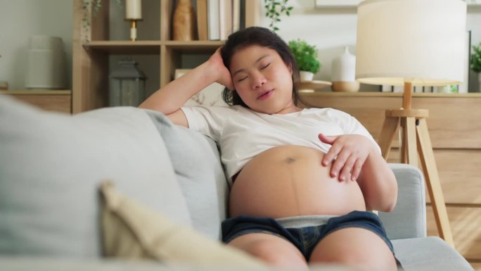 幸福微笑的亚洲成年妈妈怀孕了，坐在客厅的沙发上，温柔的抚摸着她的大肚子，快乐的妈妈检查着她的宝宝肚子