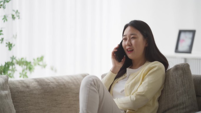 年轻的韩国女子正在打电话，美丽的亚洲女子舒适地坐在她舒适的公寓沙发上，兴奋地讨论着未来的旅行计划