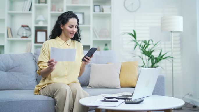 年轻女性坐在家里的沙发上，用智能手机上的网站或应用程序处理家庭财务
