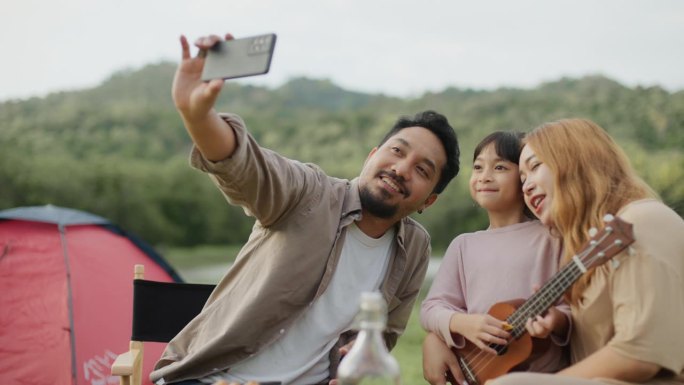 在户外露营旅行前休息的亚洲家庭。亚洲父母和女儿一起用智能手机自拍，开心的微笑享受草甸。